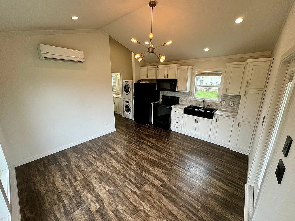 Modern kitchen with dark wood floors, white cabinets, black appliances, and a geometric chandelier. A washer and dryer are visible on the left.