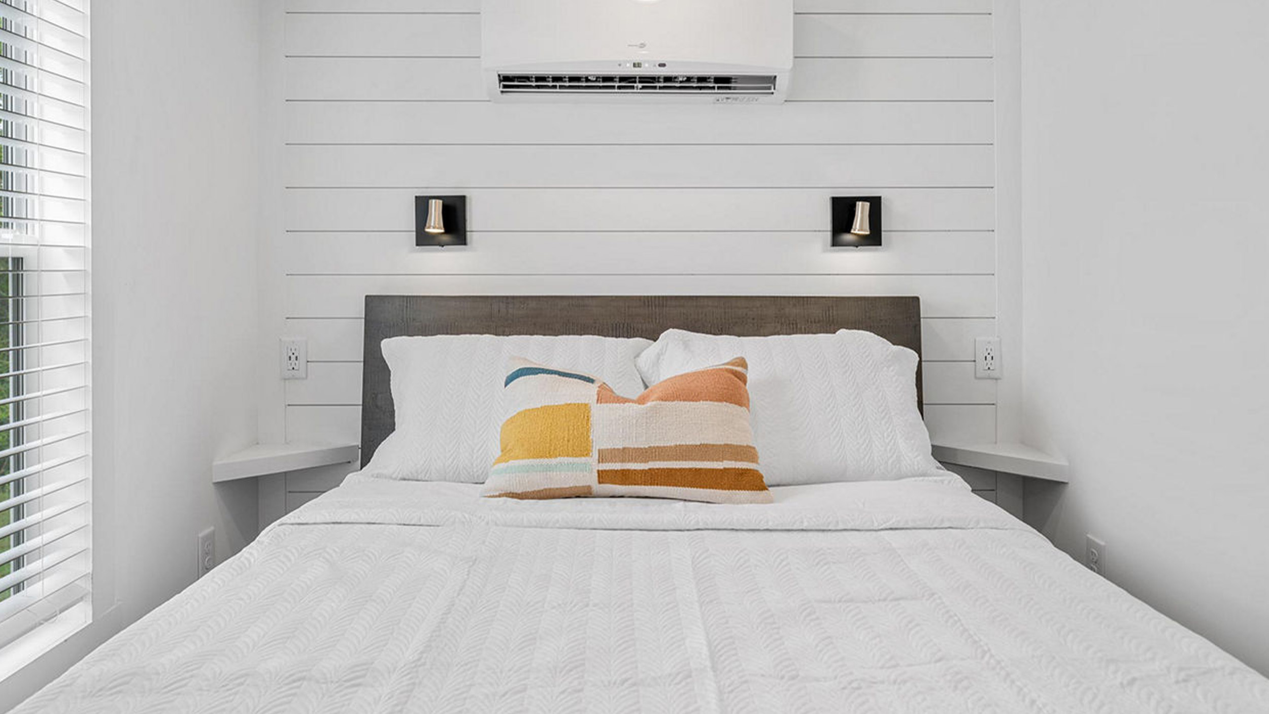 Cozy bedroom featuring a neatly made bed with white linens and colorful pillows. Soft lighting and a modern air conditioner create a comfortable atmosphere.