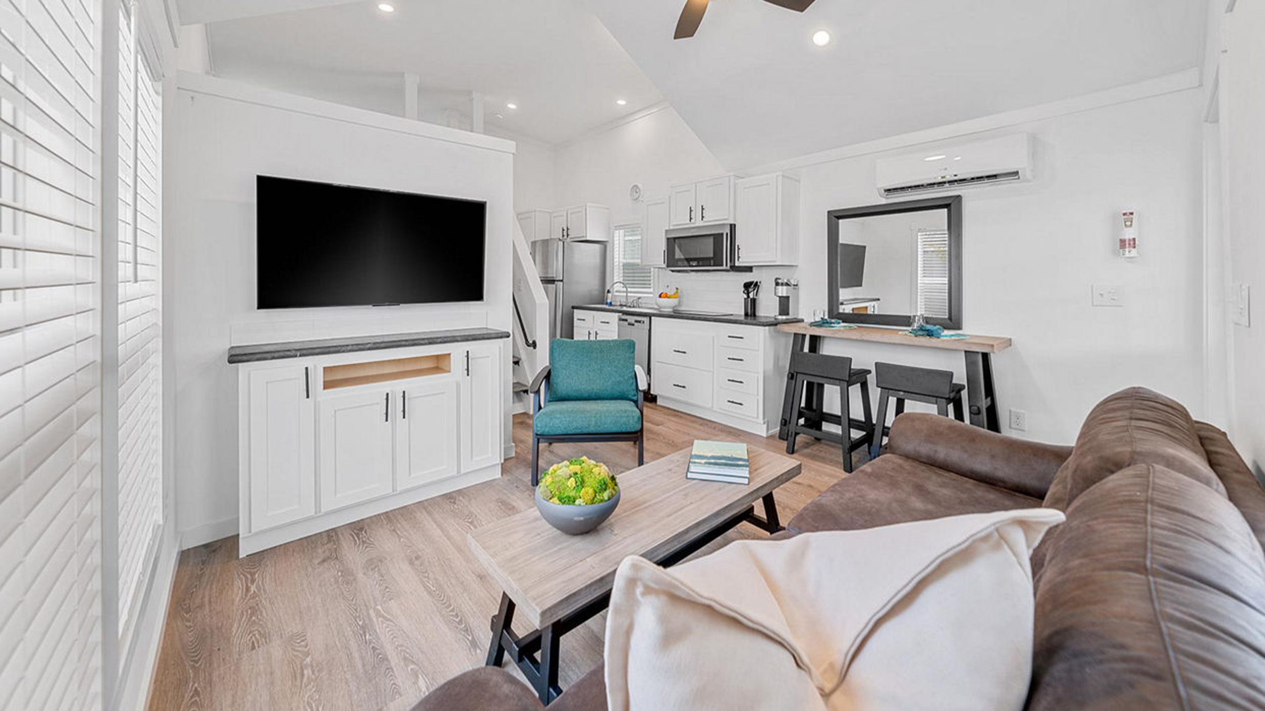 A modern, cozy living room with a brown couch, white walls, and wooden floors. A large TV on a white cabinet is opposite. The compact kitchen in the background features white cabinets and stainless steel appliances. A teal armchair and a wooden coffee table with a bowl of green apples add color. Warm, inviting ambiance.