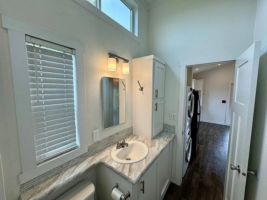 Modern bathroom with white walls, marble countertop, and a single sink. A mirrored cabinet is above the sink, flanked by two lit sconces. Wooden flooring leads to a kitchen with a stacked washer and dryer visible. The window blinds are partially closed, adding a soft illumination to the space.