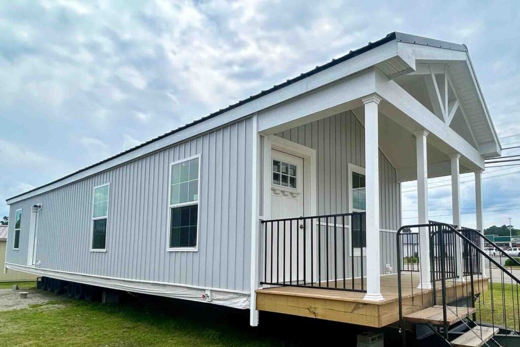 A modern tiny house with gray siding and white trimming sits on a grassy lot. It features a small wooden porch with black railings and stairs, under a cloudy sky.