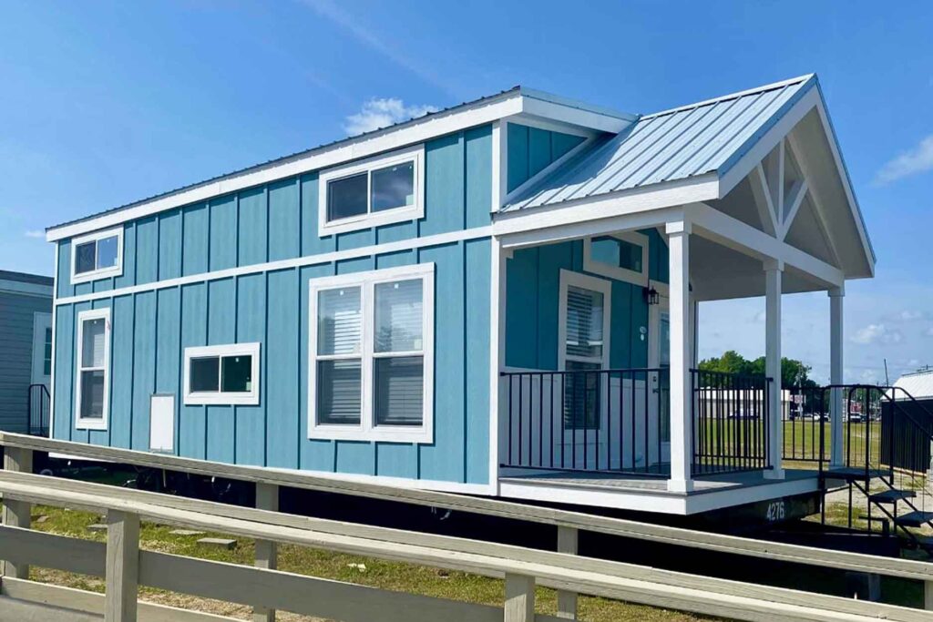 A charming blue tiny house with white trim and a gable roof is set against a clear blue sky. A welcoming front porch adds a cozy touch.