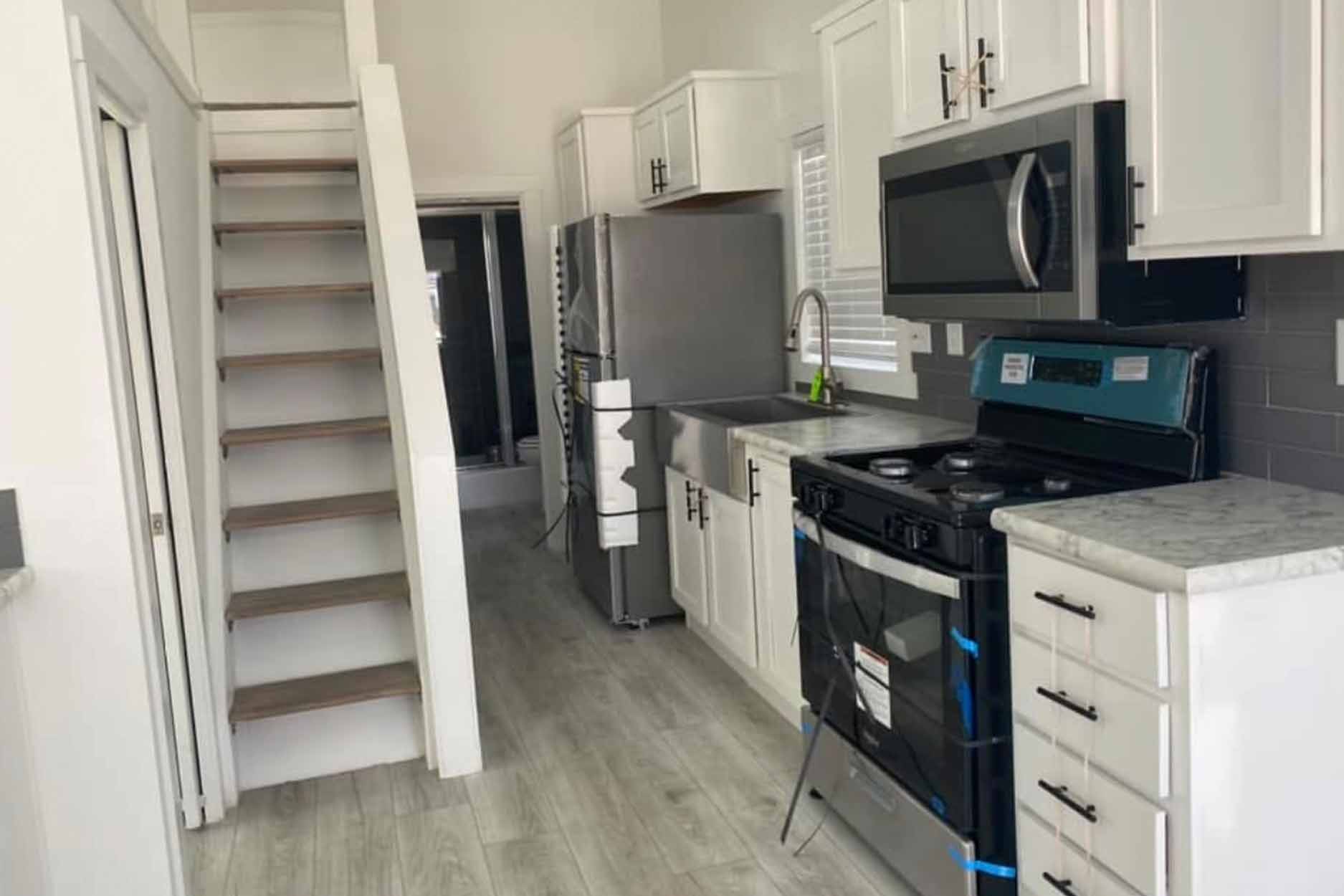 Compact modern kitchen with white cabinets, stainless steel appliances, and a gray-tiled backsplash. Wooden stairs lead to an upper level. Bright and organized space.