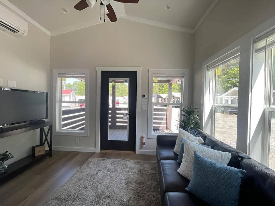 Living room with light gray walls and wooden floor, featuring a brown leather sofa with blue and white cushions. Large windows and door reveal a sunny outdoor view. A TV is mounted on a console to the left, and a ceiling fan hangs above, adding an airy, cozy atmosphere.