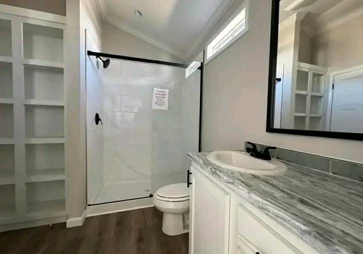 Modern bathroom with wood flooring, large shower, and white vanity with marble countertop. Shelving unit on the left. Bright and clean atmosphere.