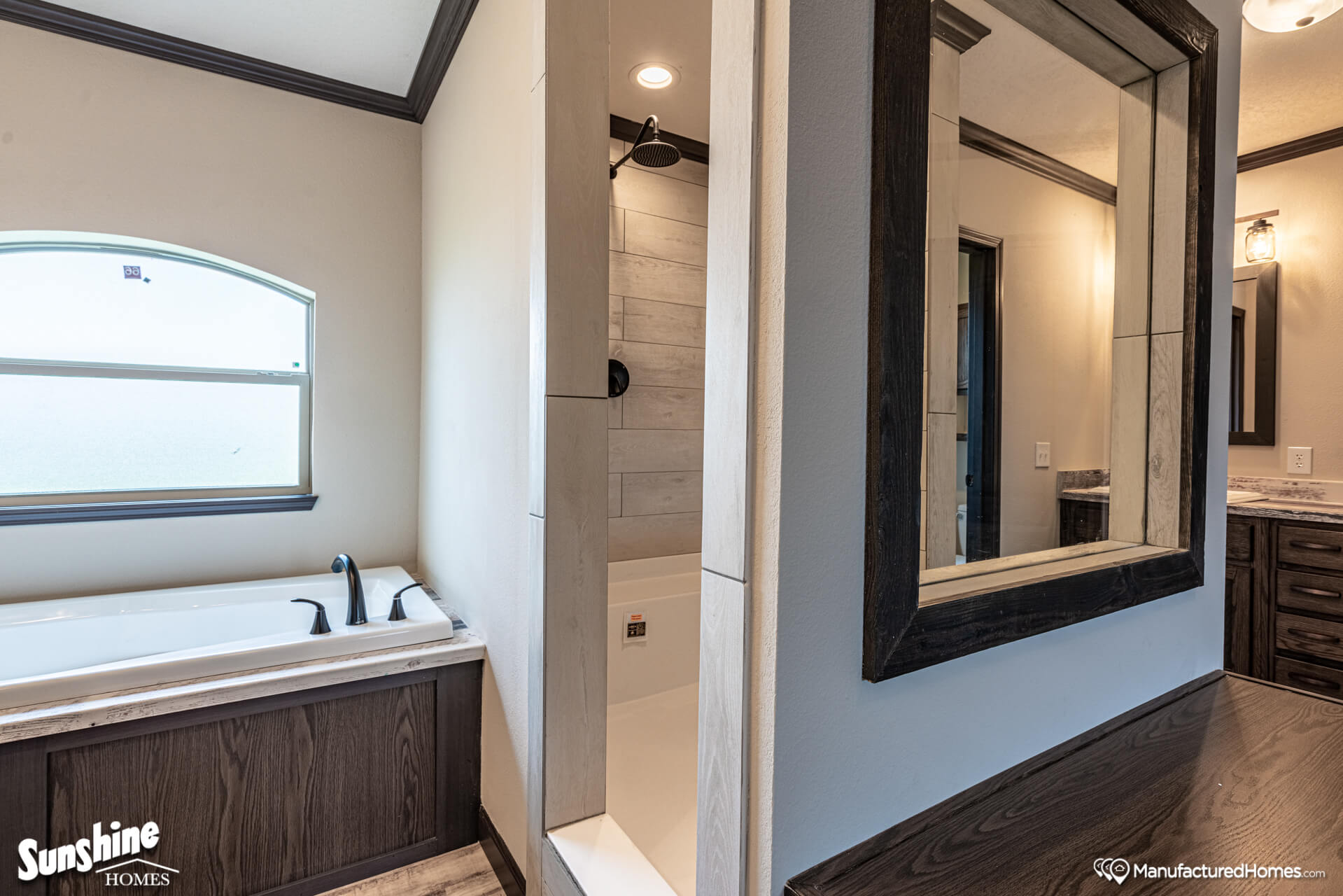 Modern bathroom with natural tones. Features a deep bathtub under an arched window, a glass-enclosed shower, and a large framed mirror on a wooden vanity.