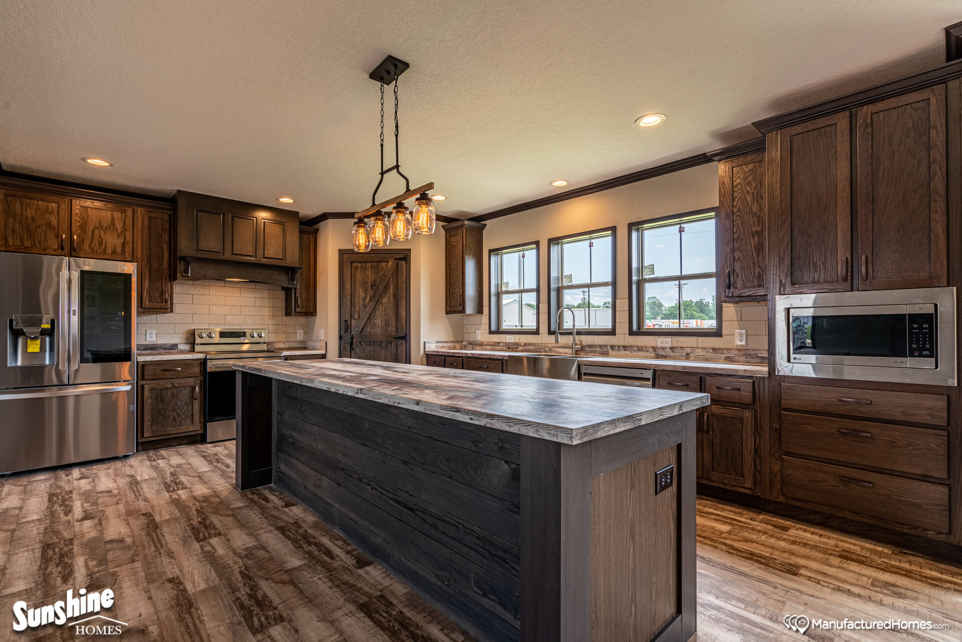 Rustic kitchen with wooden cabinets, large island, stainless steel appliances, and modern light fixture. Warm tones create a cozy, inviting atmosphere.