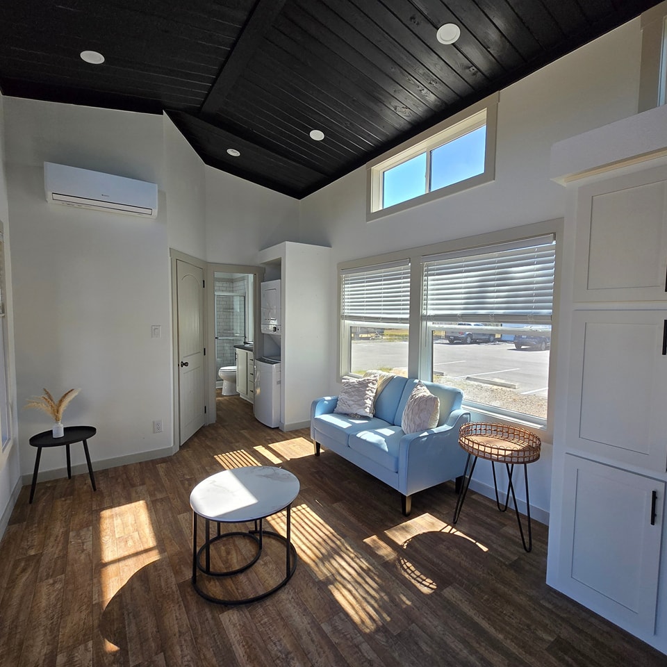 Bright modern living room with black ceiling, white walls, and wood floor. Features a light blue sofa, round coffee tables, and large windows. Cozy and airy atmosphere.