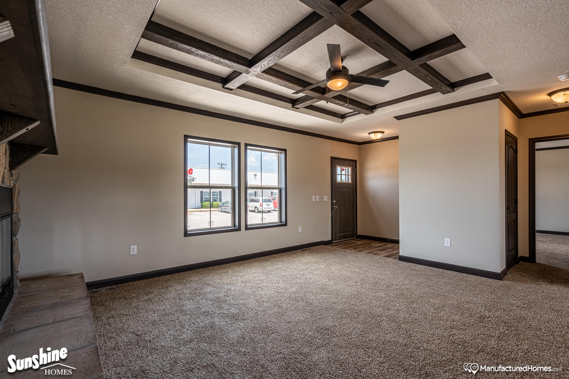 Modern living room with beige carpet, coffered ceiling, and ceiling fan. Large windows provide natural light. Warm, inviting atmosphere with textured walls.