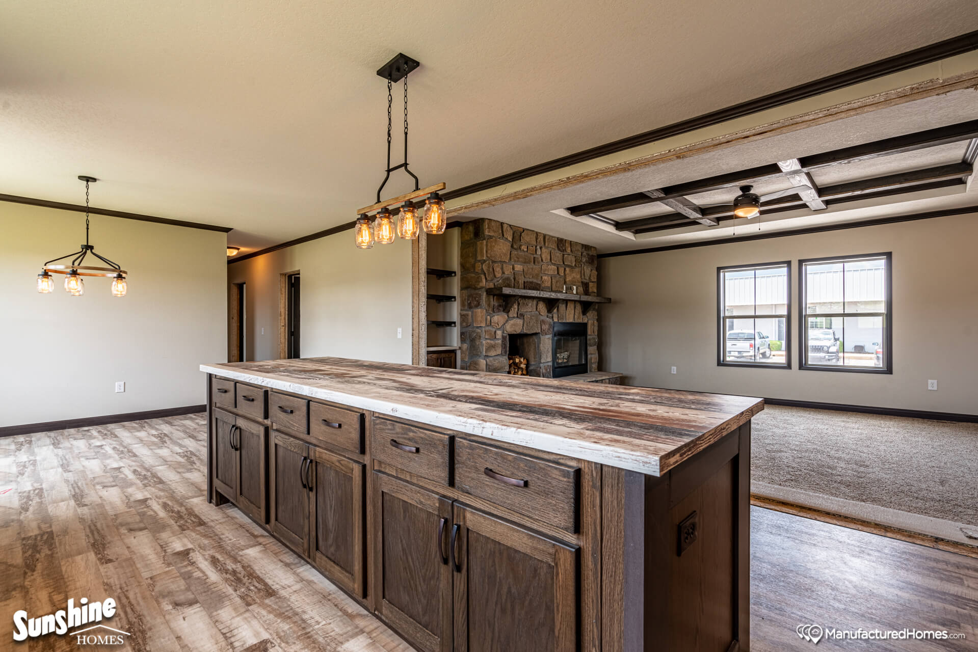 Spacious open-plan living area with a rustic kitchen island, modern lights, stone fireplace, and large windows. Warm, inviting atmosphere.