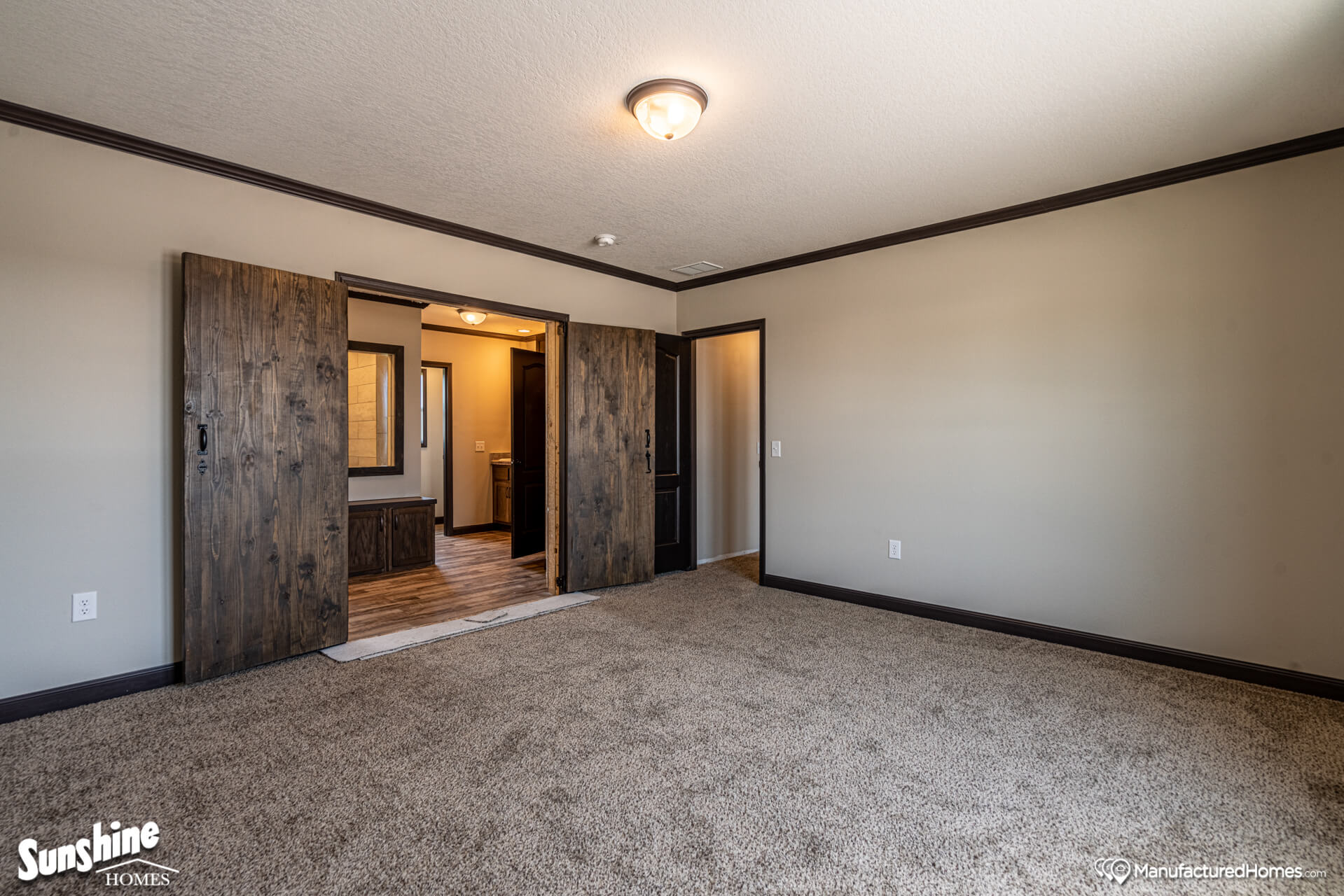 Spacious room with beige walls, carpeted floor, rustic wooden sliding doors, ceiling light, and view into an adjacent room with wood flooring. Cozy ambiance.