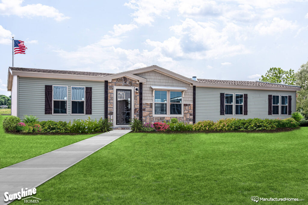 A single-story manufactured home with light gray siding and stone accents, surrounded by a lush green lawn and a walkway. An American flag flies to the left.