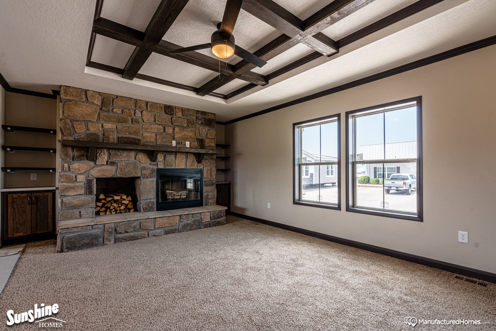 Cozy living room with beige carpet, stone fireplace, ceiling fan, and wooden beam ceiling. Bright windows offer outdoor views, creating a warm ambiance.