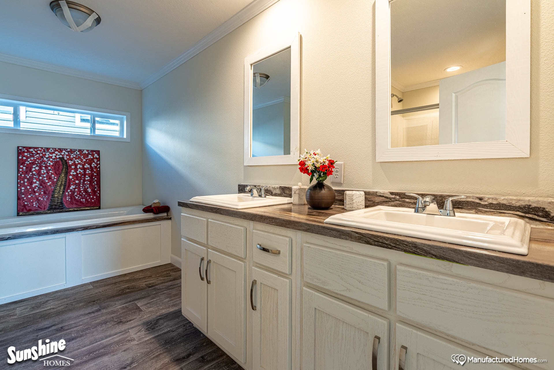 Bright bathroom featuring dual sinks with mirrors, white cabinets, and a vase of red flowers. Artwork adorns the wall, and light streams through a high window.