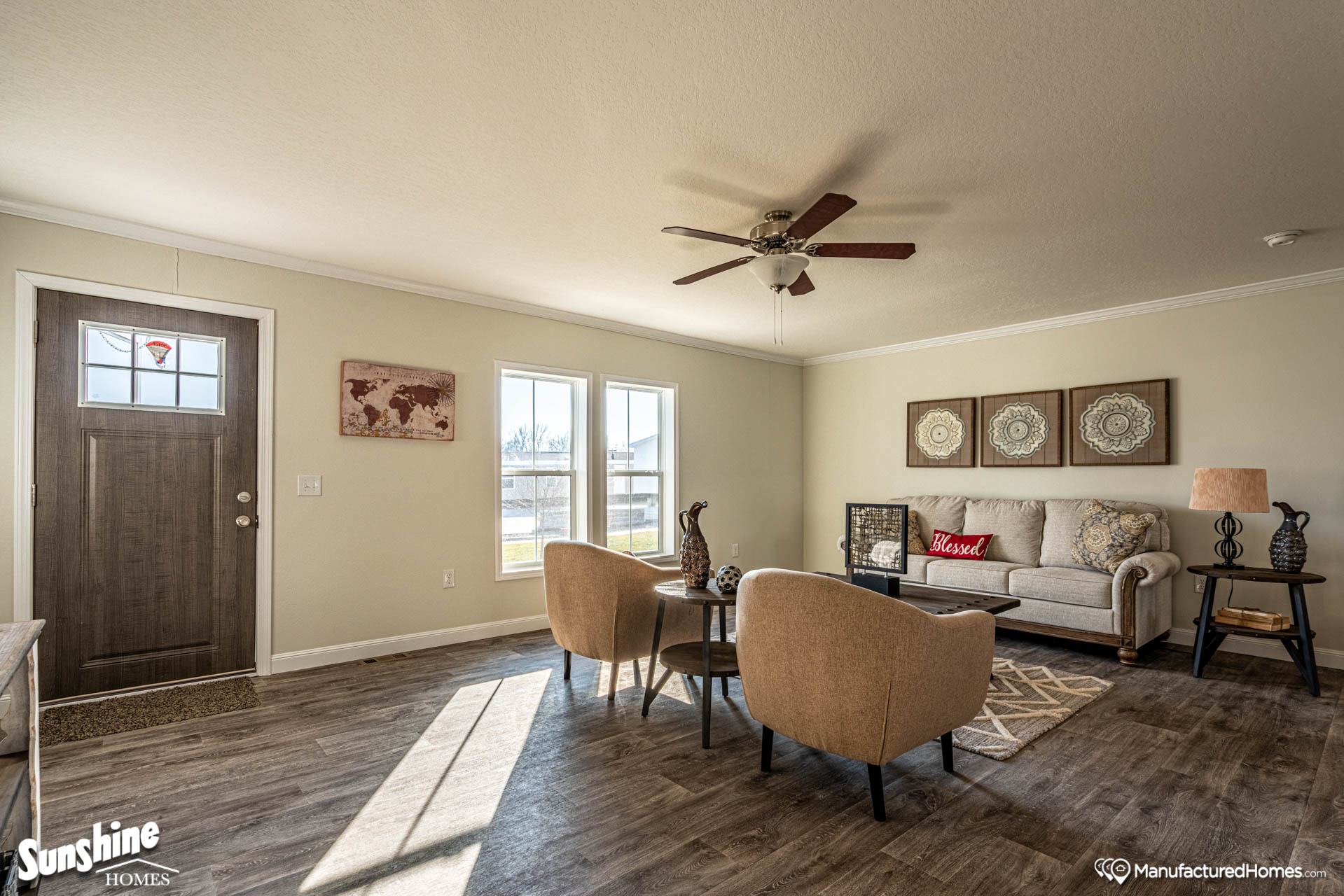 Bright living room with wood flooring, a ceiling fan, and natural light from large windows. Cozy seating area with beige chairs and a sofa, accented by pillows and wall art. Warm, inviting atmosphere.