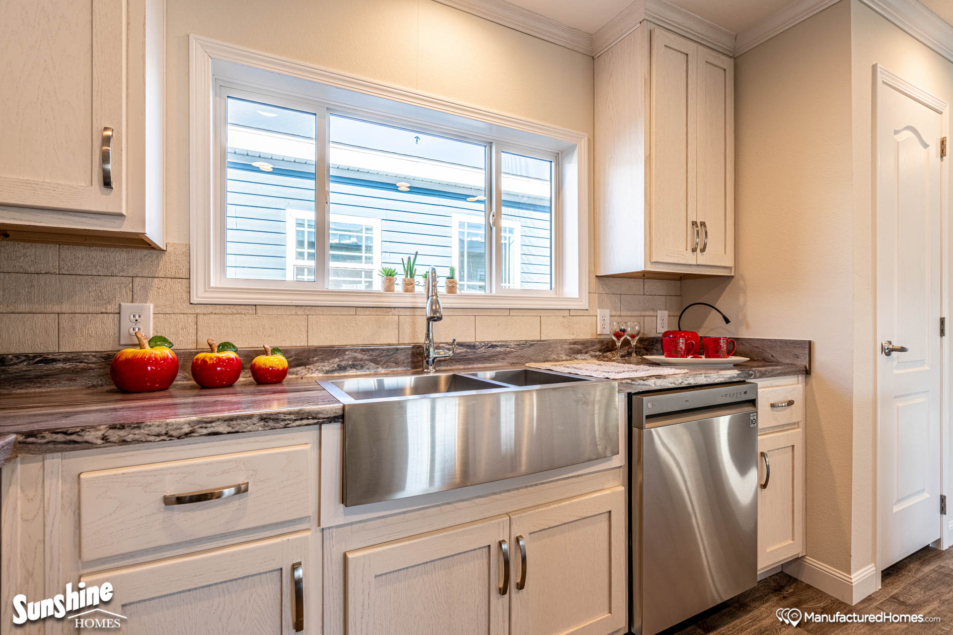 Modern kitchen with a stainless steel farmhouse sink, white cabinets, and gray countertops. Red apples and a plant adorn the space, creating a fresh, inviting atmosphere.
