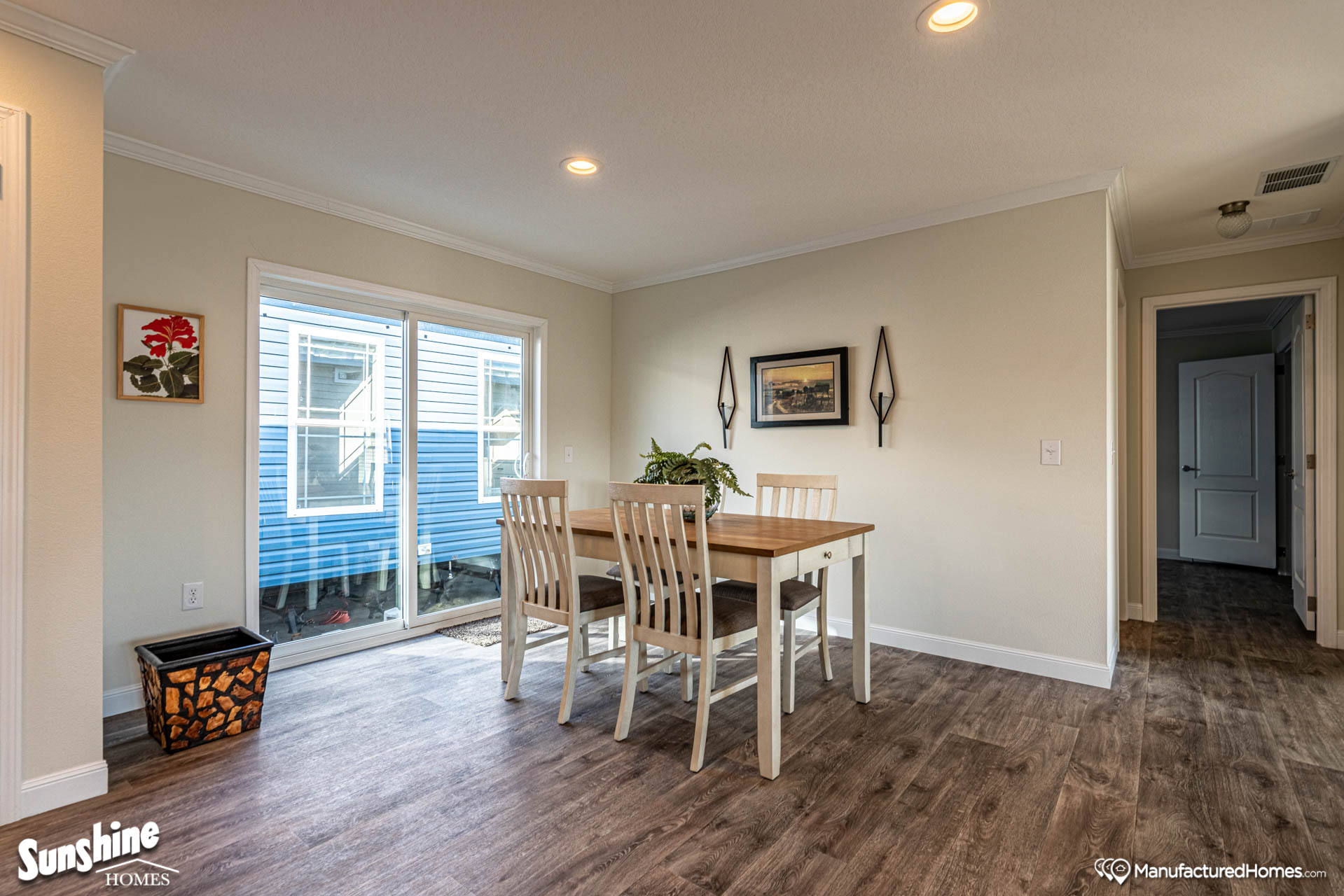 Bright dining room with wooden floors features a table and four chairs near sliding glass doors. Wall decor includes a painting and sconces, creating a welcoming atmosphere.