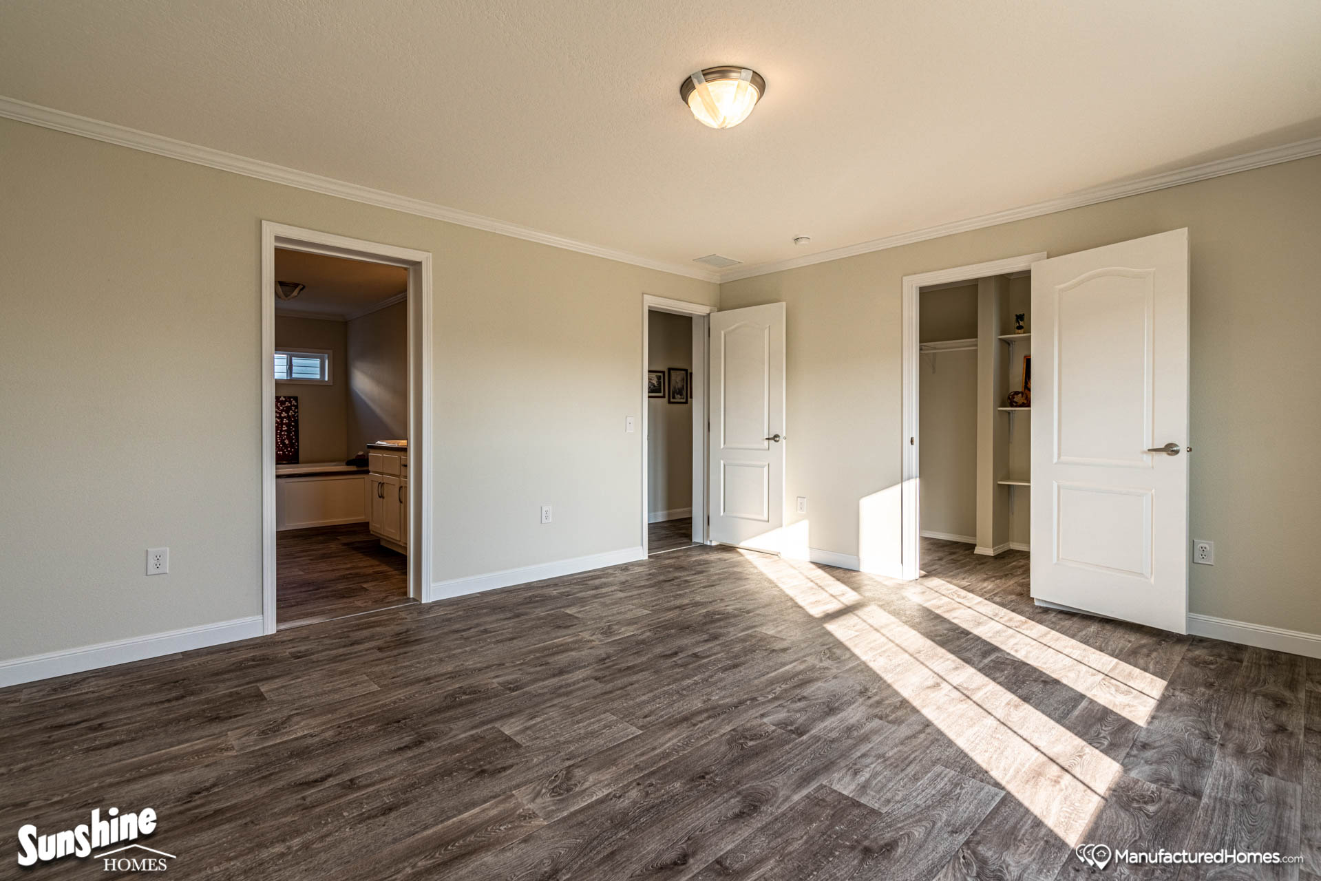 An empty room with light beige walls and wood-look flooring has two open doorways, one leading to a bathroom and the other to a closet. Sunlight streams in, creating a warm, inviting atmosphere.
