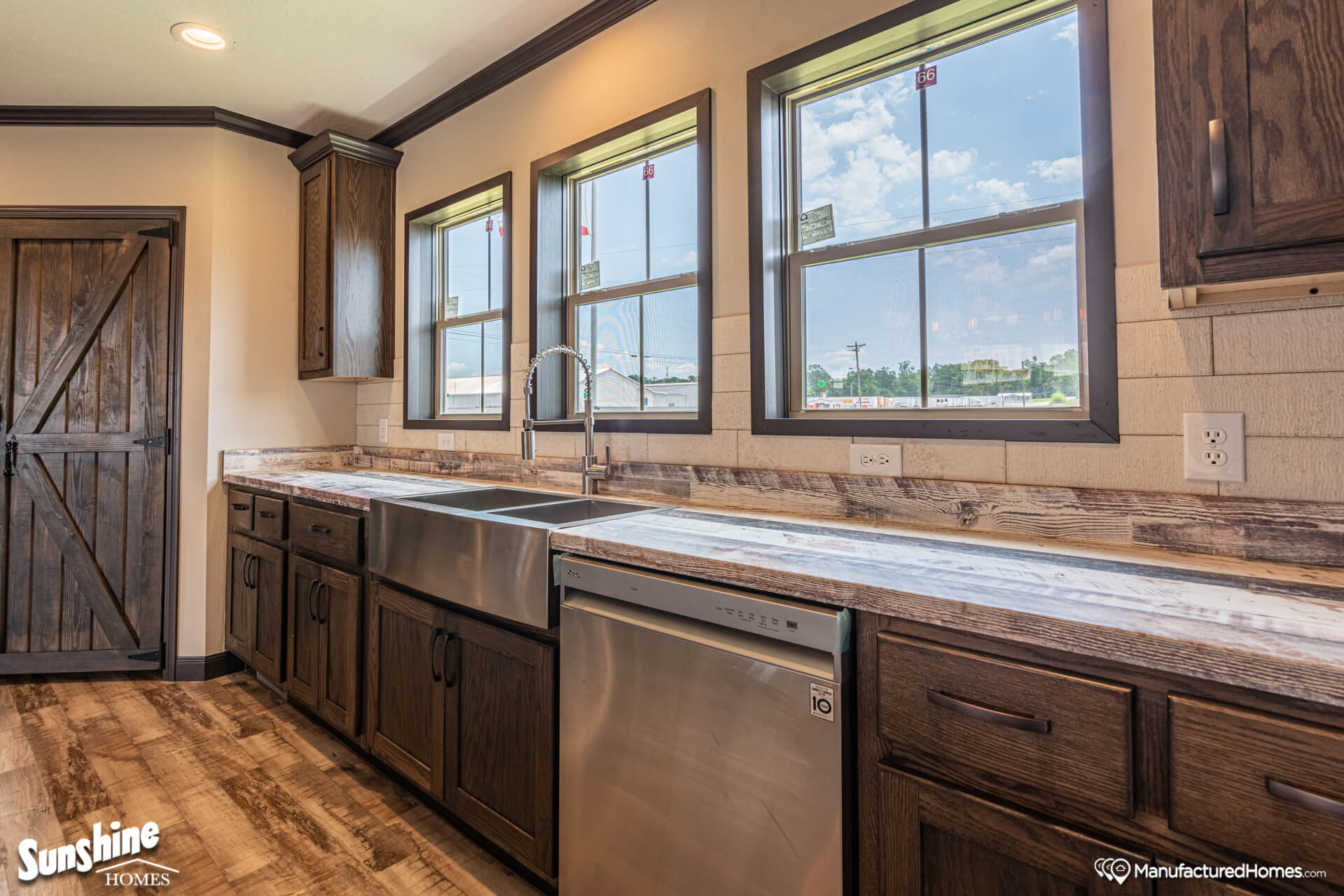 Rustic kitchen with dark wood cabinets, stainless steel dishwasher and double sink. Three windows above the sink fill the room with natural light.