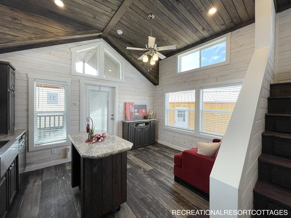 Modern tiny home interior with vaulted wooden ceiling, large windows, and dark wood flooring. Features a small kitchen, red sofa, and loft stairs. Cozy ambiance.