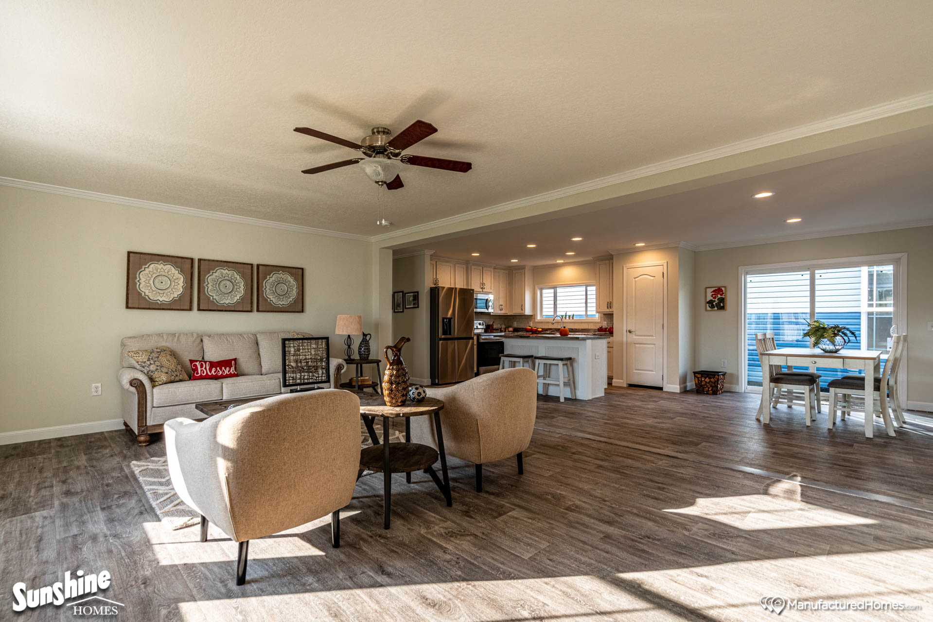 Spacious, cozy living room with tan armchairs, a couch with "Blessed" pillow, and a ceiling fan. Open kitchen and dining area with wooden floors.