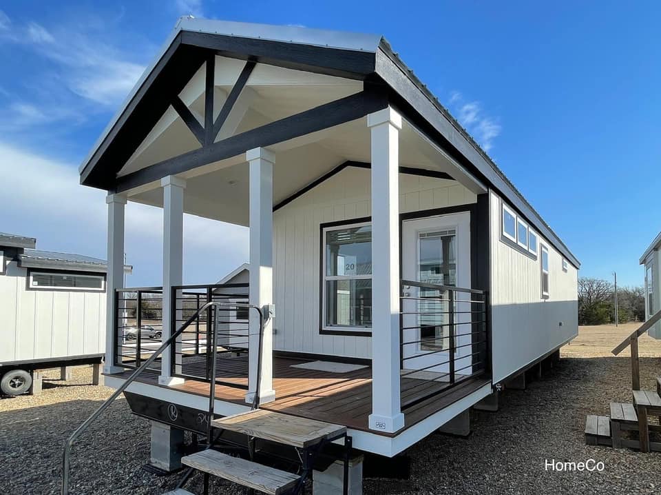 Modern tiny house with black and white trim, front porch, and stairs, set against a blue sky. The setting is open and sunny, creating a welcoming ambiance.