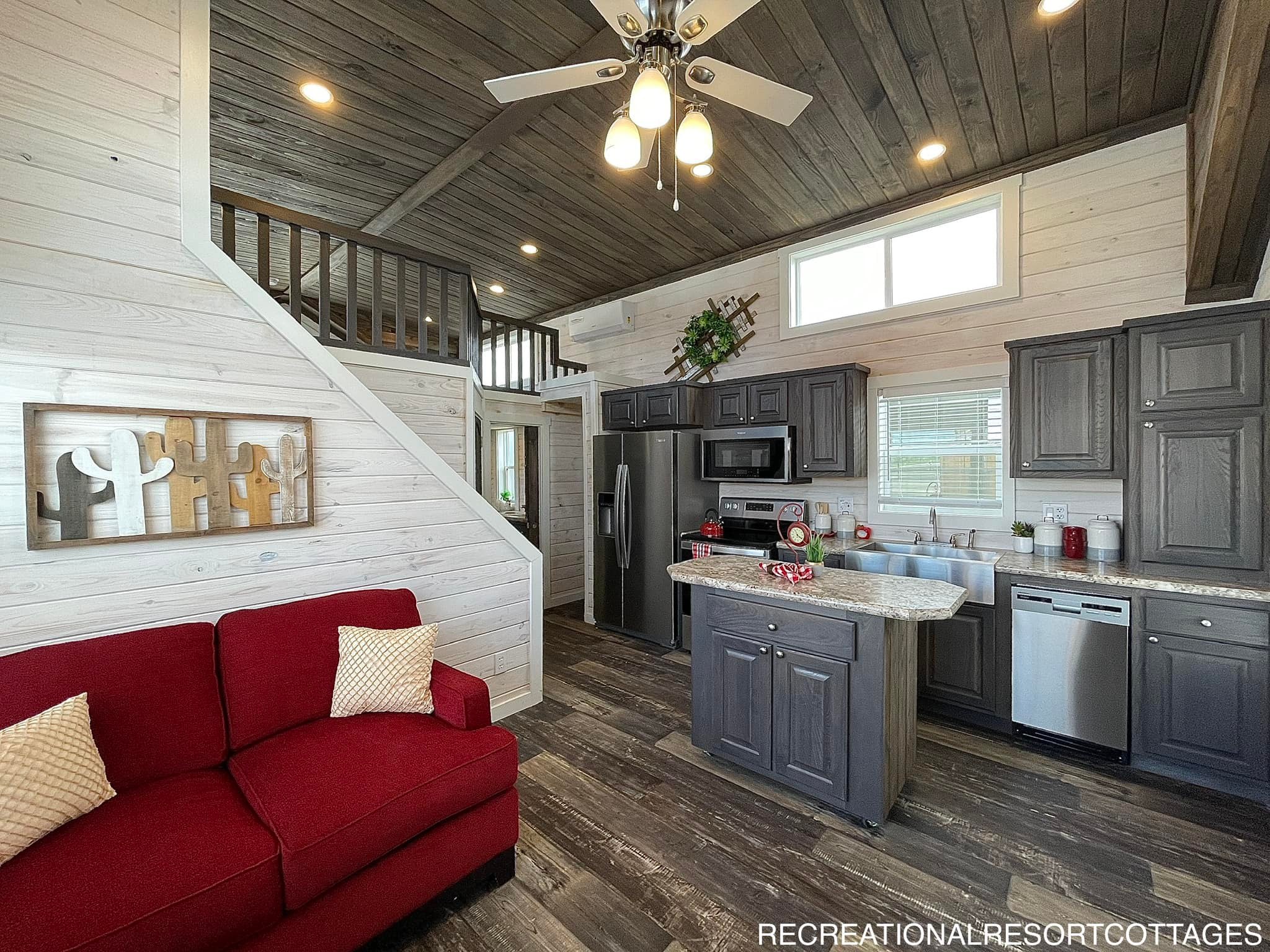 Cozy interior of a small home with rustic wooden accents. Features a red sofa, sleek modern kitchen, loft area, and ceiling fan, creating a warm ambiance.