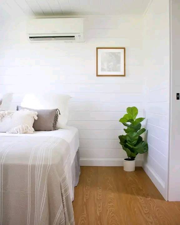 Bright minimalist bedroom with white walls and bedding. A potted plant adds freshness, and a framed picture hangs above. An air conditioner is mounted high.