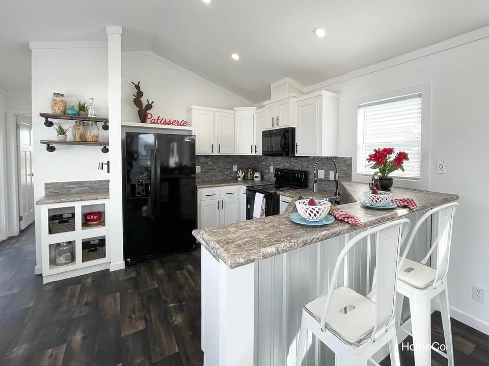 Modern kitchen with white cabinets, dark appliances, and marble countertops. A breakfast bar with white stools holds dishes and flowers, creating a cozy feel.