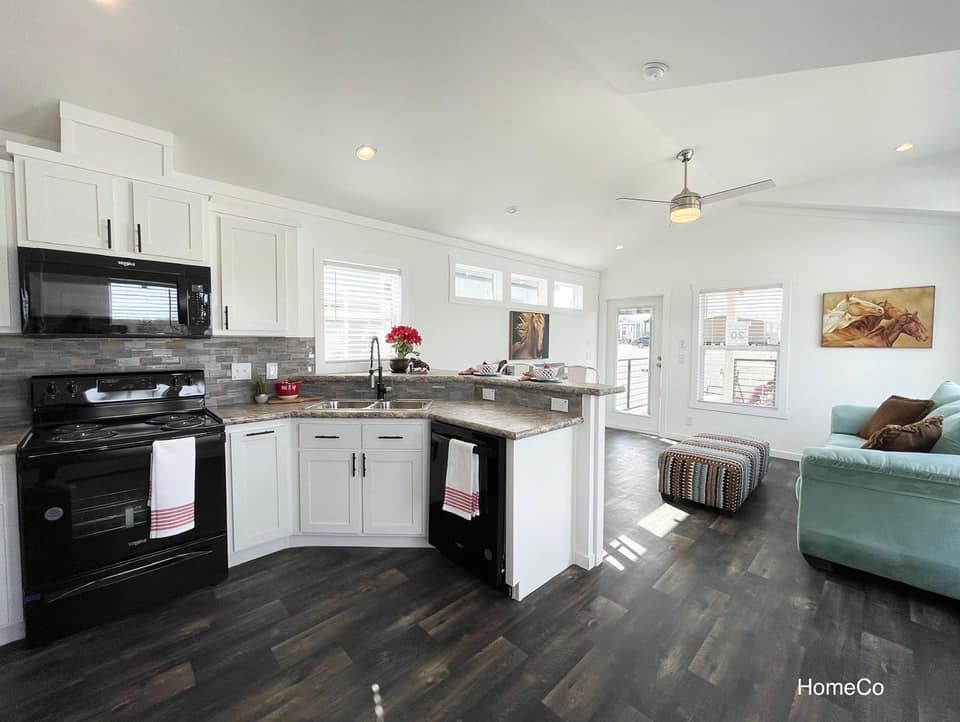 Modern open kitchen and living area with dark wood flooring. Features white cabinets, black appliances, a teal sofa, and natural light, conveying a cozy, airy feel.
