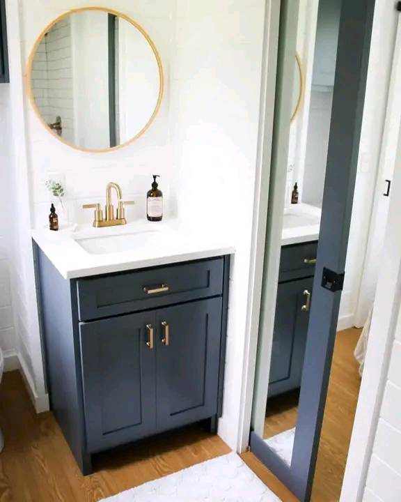 Modern bathroom with a gray vanity, white countertop, brass faucet, and round mirror. A sliding mirror door and wood floor create a clean, sleek vibe.