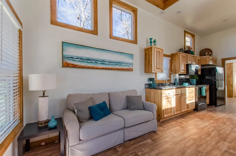Bright living room and kitchen with wooden floors and cabinets. Gray sofa with blue cushions, coastal artwork, and large windows. Calm and cozy atmosphere.