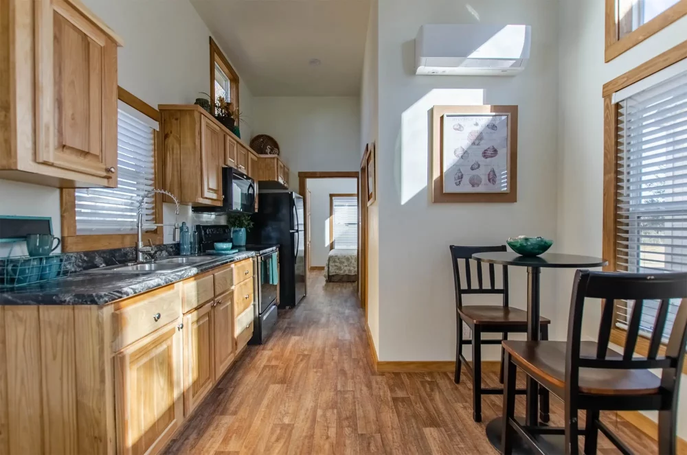 Modern kitchen with wooden cabinets and flooring, black appliances, and a narrow hallway leading to a sunlit bedroom. Cozy dining nook on the right.