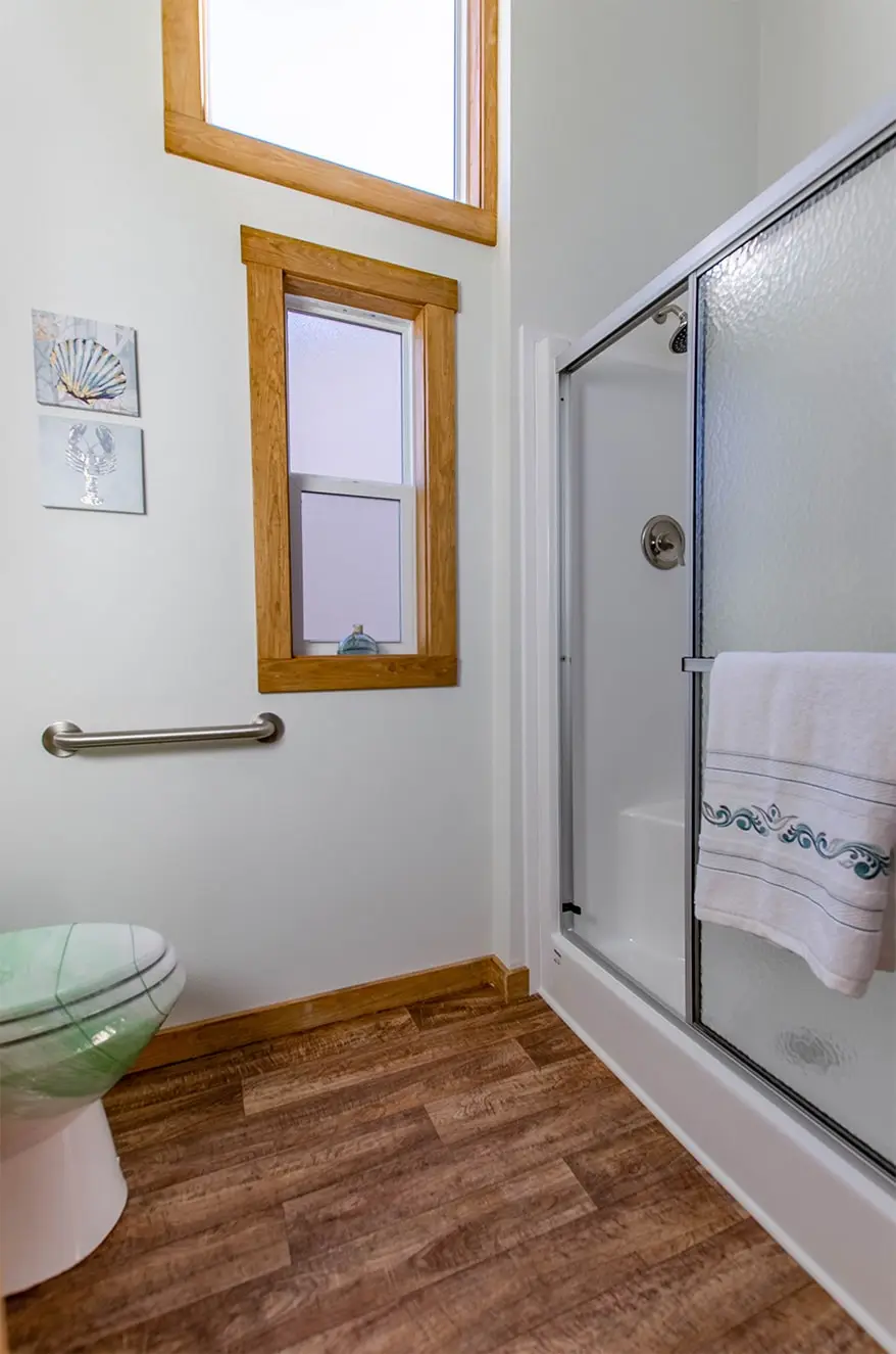 Bright bathroom with wood-trimmed windows and vinyl wood flooring. Features a shower with a glass door, a towel bar, and coastal decor on the wall.
