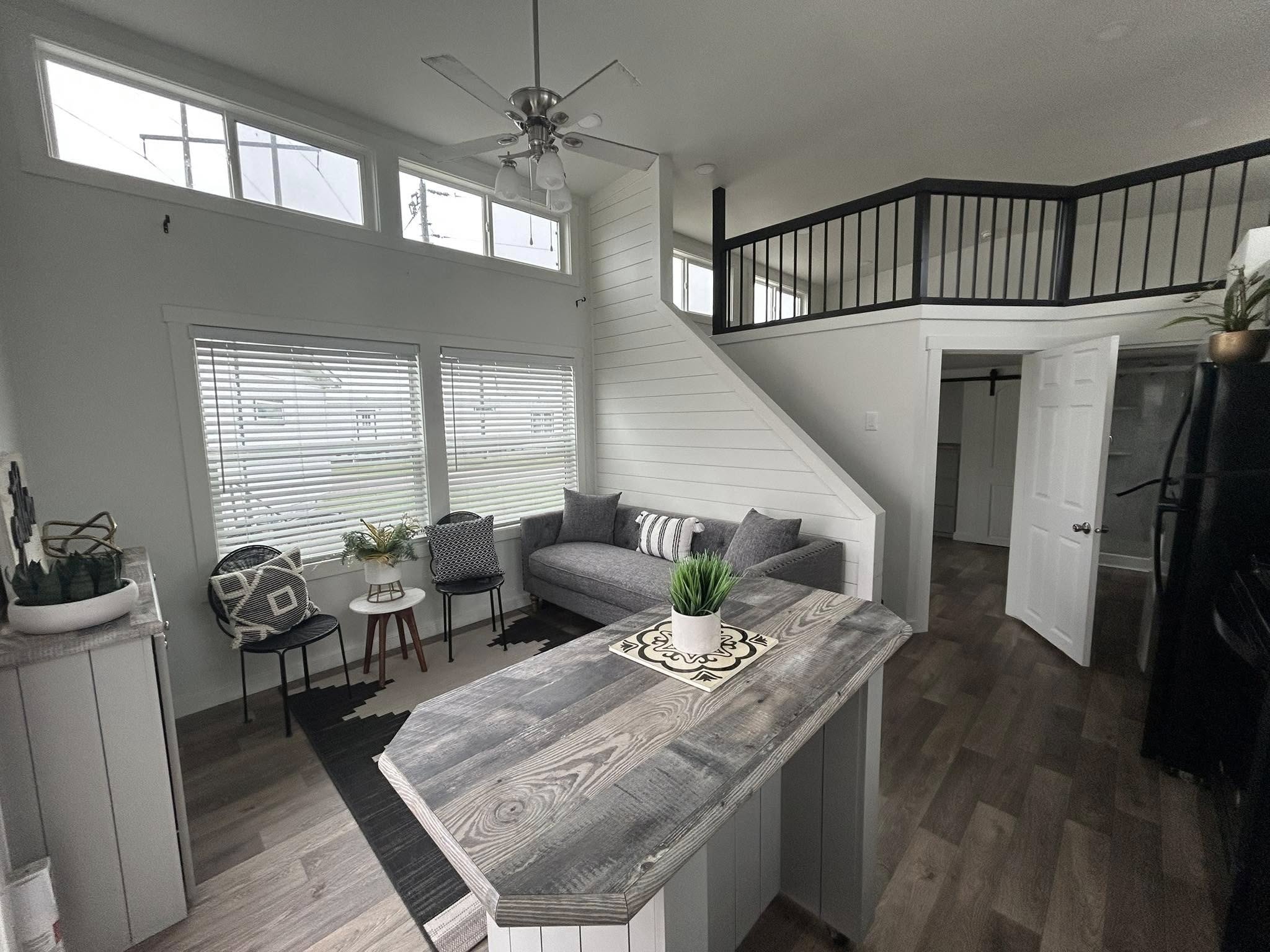 This diverse collection of luxury tiny homes, park models, and manufactured homes highlights the signature "The Boujee XL" aesthetic, blending rustic charm with high-end modern finishes. Exterior architectural styles feature cream-colored board-and-batten siding with white metal roofs and black-pillared porches, as well as a rustic log-sided cabin model with a green metal roof and a spacious gabled front porch. The interiors emphasize a "modern boujee" lifestyle, showcasing vaulted ceilings with exposed natural wood beams, white shiplap walls, and integrated lofted sleeping areas with black metal or wooden railings. Kitchen designs offer a range of styles, from rustic wood cabinetry with stone-look countertops to sophisticated grey-toned cabinetry with stainless steel farmhouse sinks and distressed wood islands. Functional luxury is consistent throughout the open-concept living spaces, featuring wide-plank wood-look flooring, industrial-style ceiling fans, and large transom windows that flood the rooms with natural light.