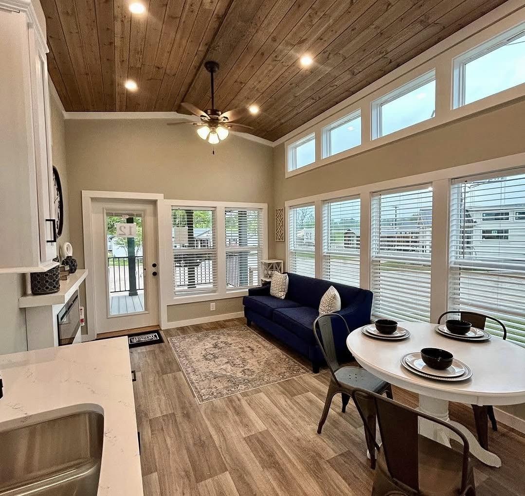 Cozy living space with a wooden ceiling, ceiling fan, and large windows. Blue sofa, round dining table set, and neutral tones create a warm, inviting feel.