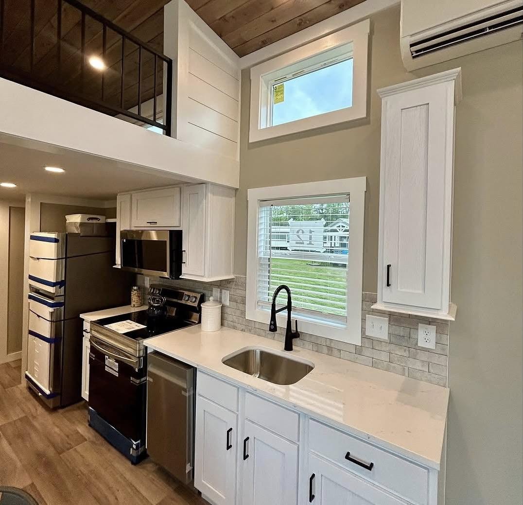 Compact modern kitchen with white cabinets, stainless steel appliances, and black faucet. Wooden ceiling and bright windows create a cozy ambiance.