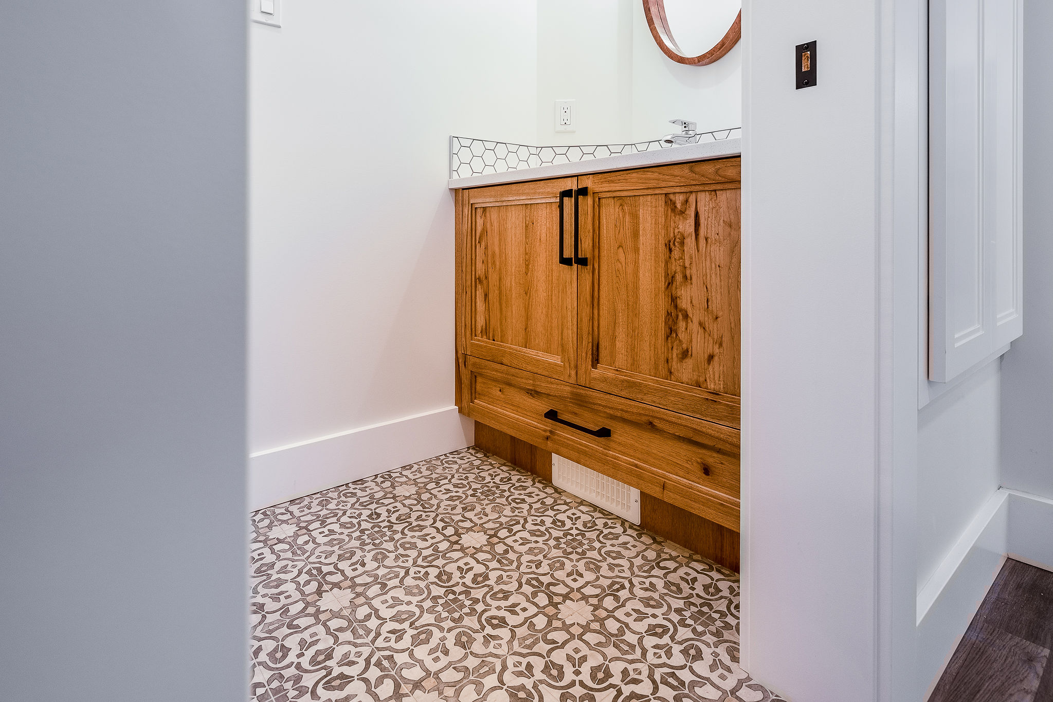 A small bathroom with a wooden vanity and black handles, patterned tile flooring, and a minimalist white countertop with a round mirror above.