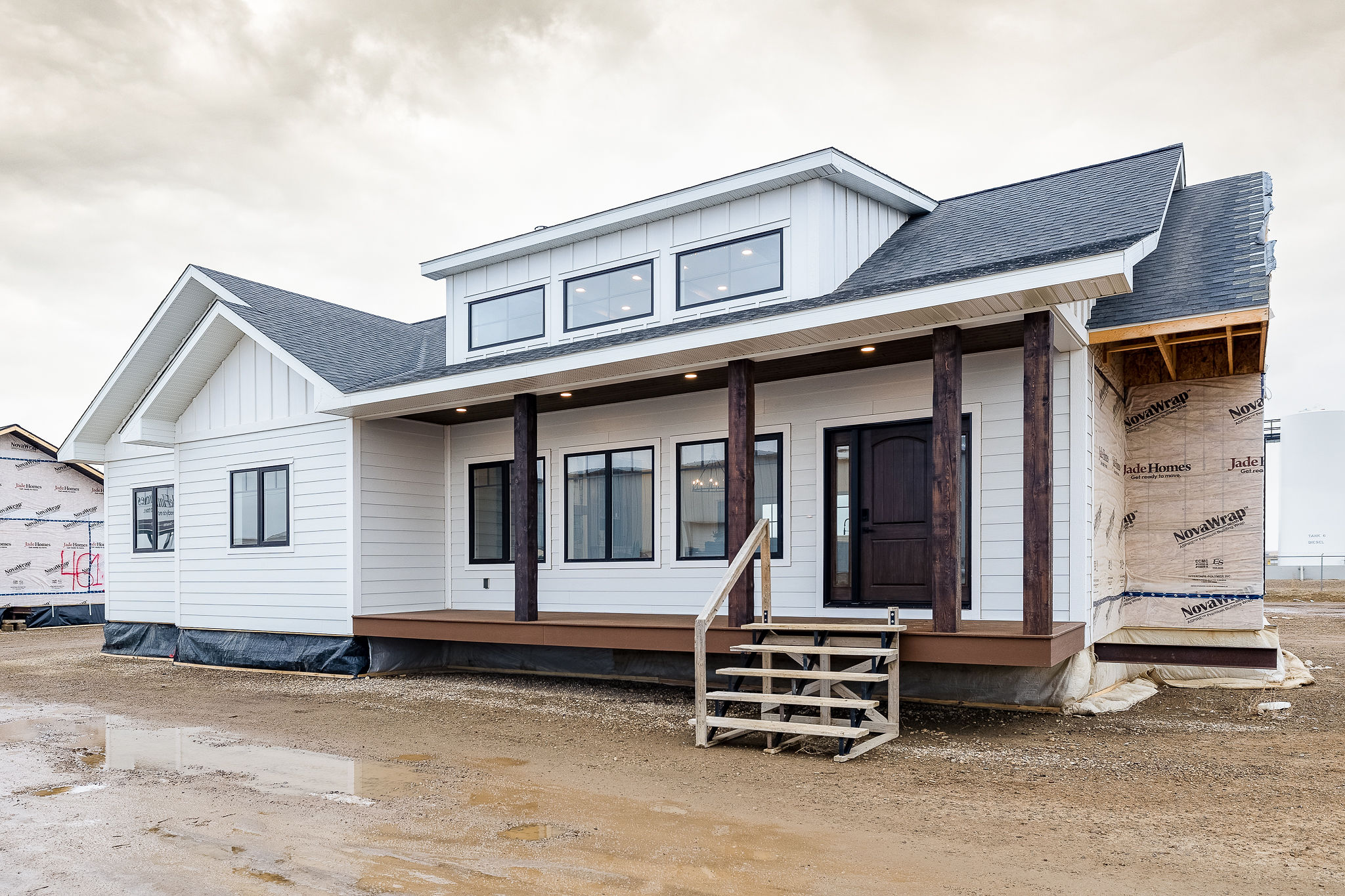 Modern white prefab house with dark wooden accents, steps leading to a porch, and a partially constructed extension. Overcast sky with soft, diffused light.
