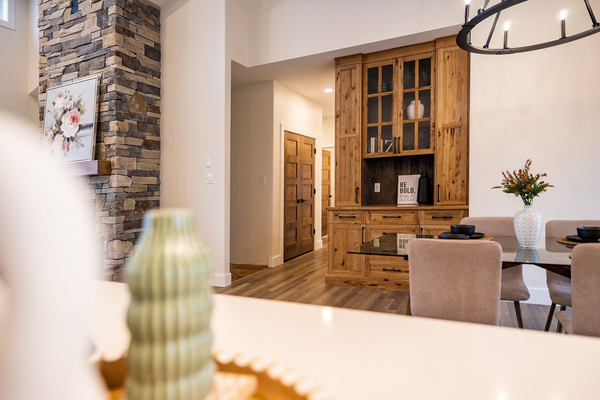 Cozy dining area with a wooden cabinet, glass table, and beige chairs. A stone fireplace with floral art adds warmth. Modern decor, calming ambiance.