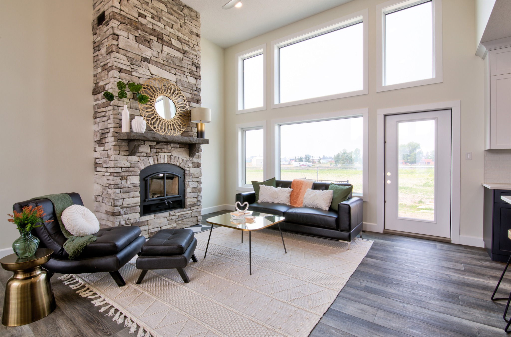 A modern living room features a tall stone fireplace, black leather chairs, and sofa with colorful cushions. Large windows provide abundant natural light.
