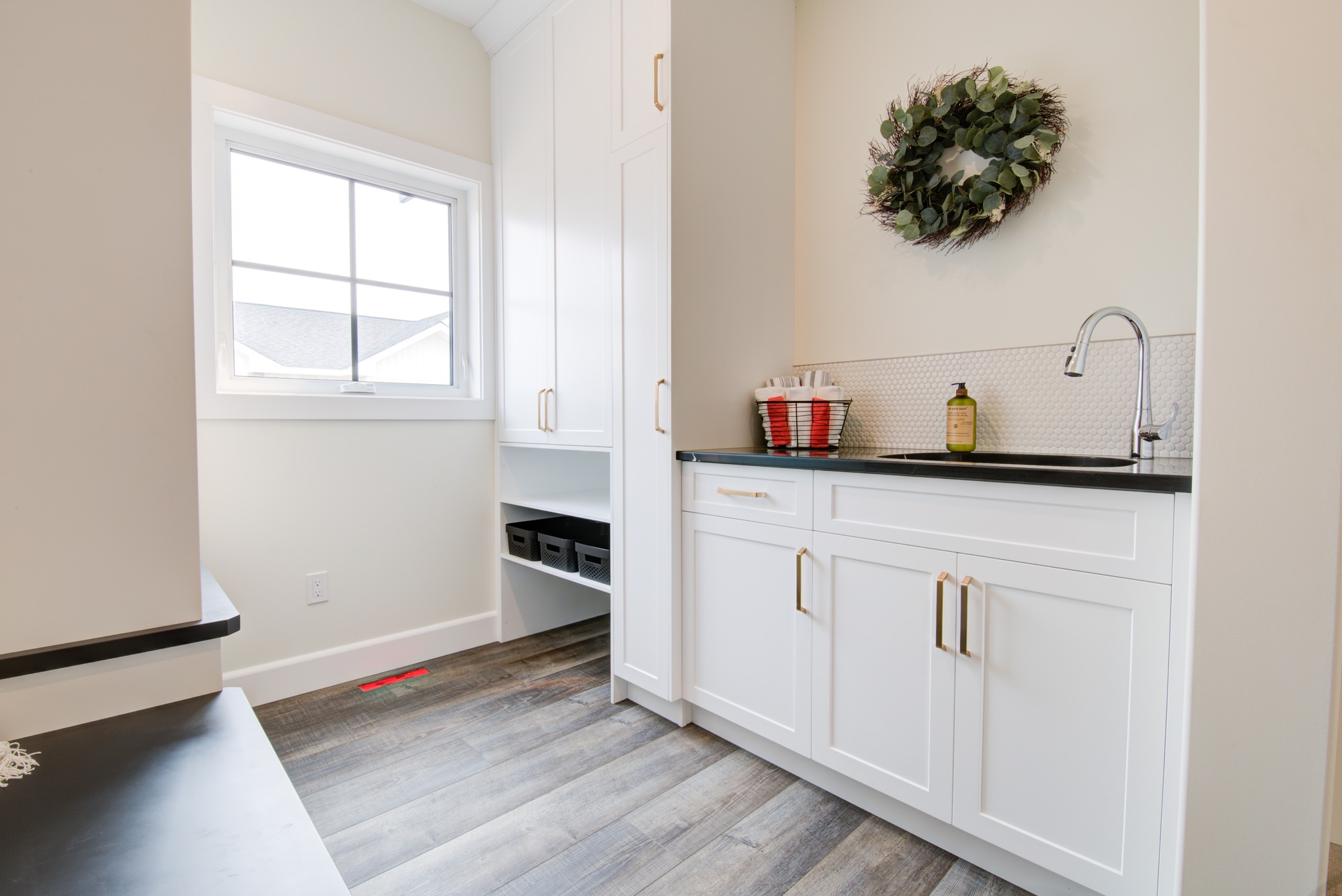 Bright laundry room with white cabinets, a black countertop, a sleek faucet, and a green wreath on the wall. A window lets in natural light. Cozy and organized.
