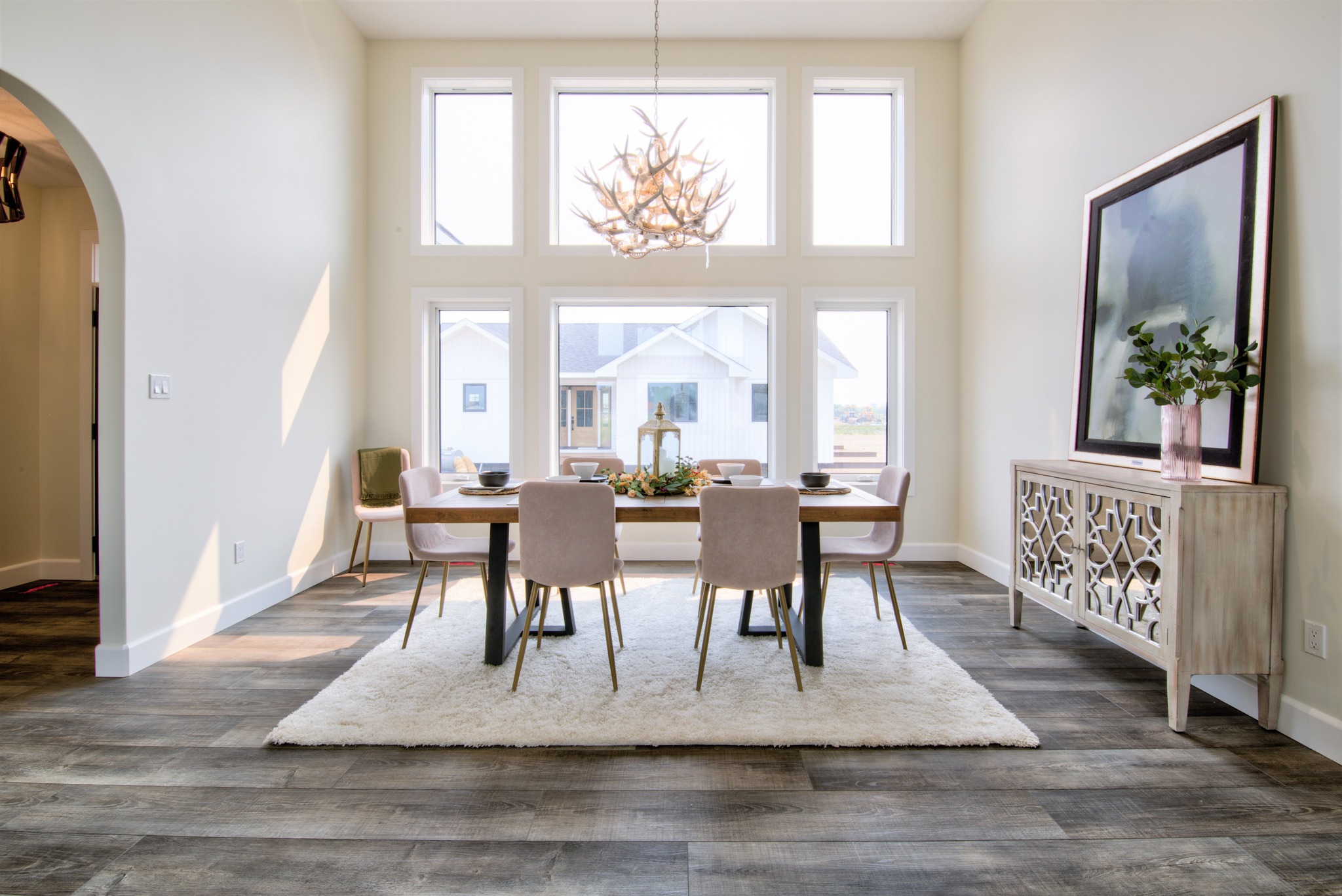 Modern dining room with large windows, a wooden table set for six, and a unique chandelier. Light wood floors and a decorative sideboard add elegance.