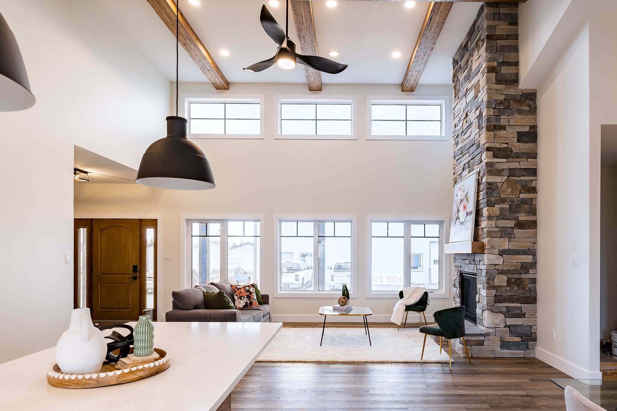 Modern living room with high ceiling, exposed wooden beams, and large windows. Features a stone fireplace, sleek furnishings, and warm lighting.