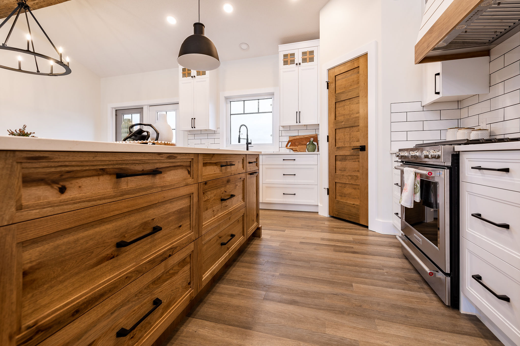 Modern kitchen with a rustic touch, featuring light wood flooring, wooden cabinets, white backsplash, black pendant lights, and stainless steel appliances.