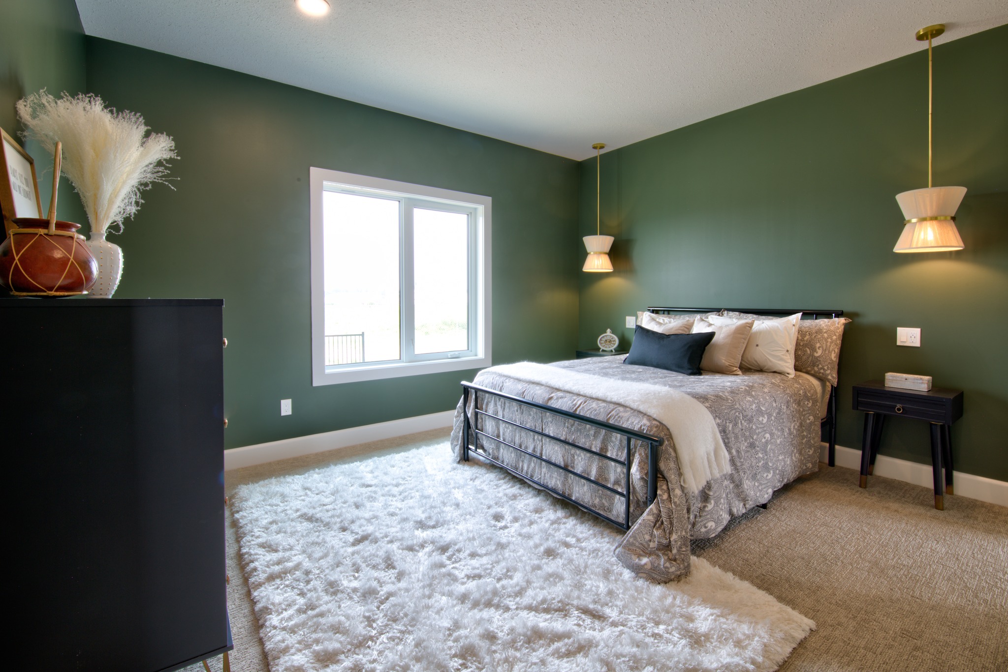 A cozy bedroom with green walls, featuring a metal bed with gray and beige bedding, soft white rug, pendant lights, and a large window for natural light.