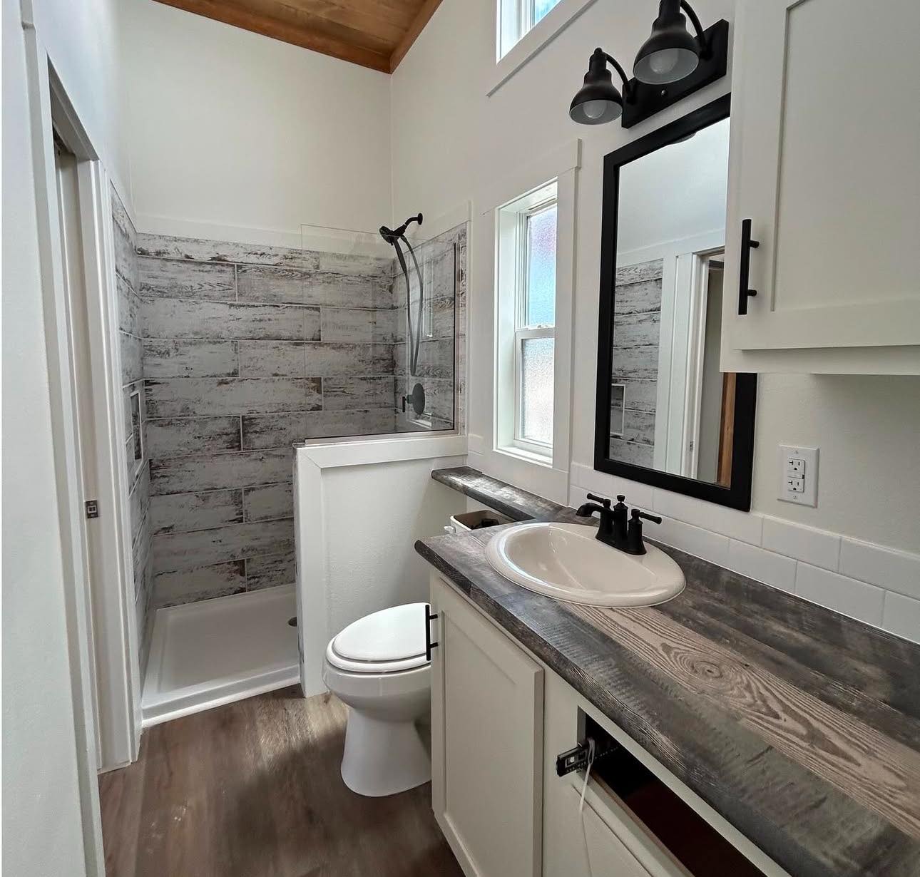Small modern bathroom with rustic wood accents. Features a shower, toilet, and sink with dark faucet. Light gray tiles and natural light create a cozy atmosphere.