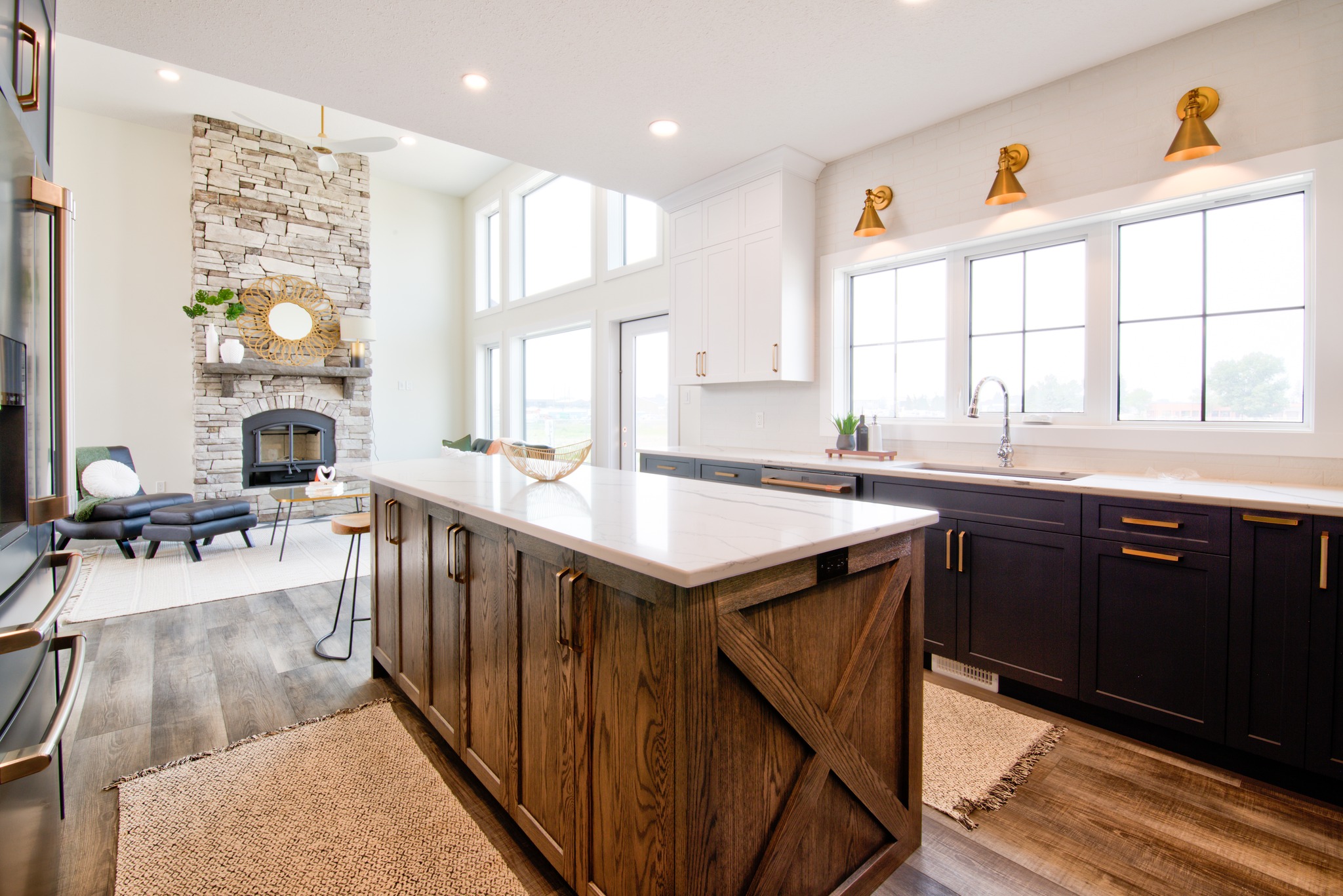 Modern kitchen with large island, stone fireplace, and high windows. White cabinets, dark lower units, warm lighting; bright, inviting space.