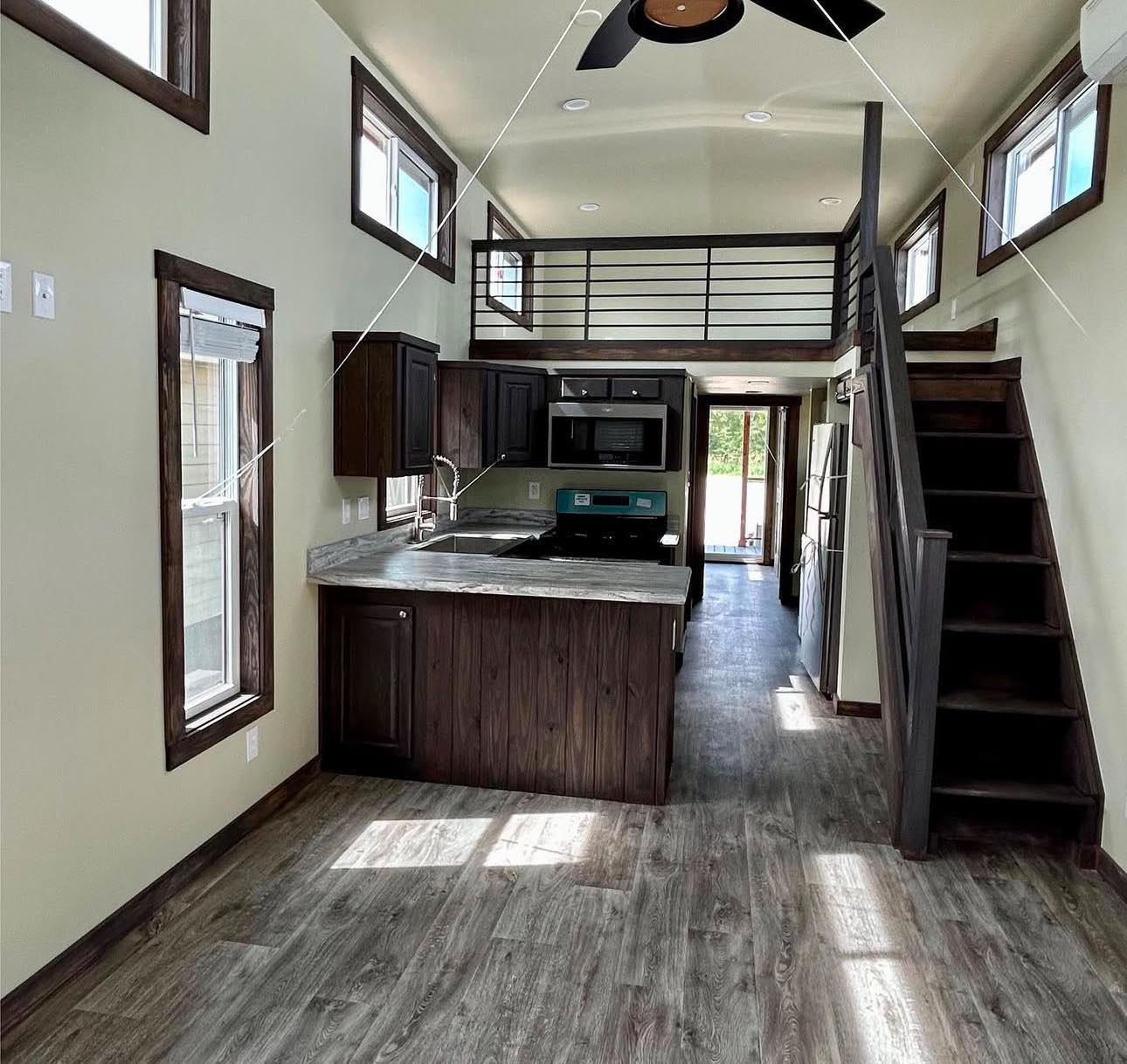 Modern tiny home interior with wood accents and open layout. Features loft, compact kitchen, stairs, ceiling fan, and natural light from windows. Cozy ambiance.
