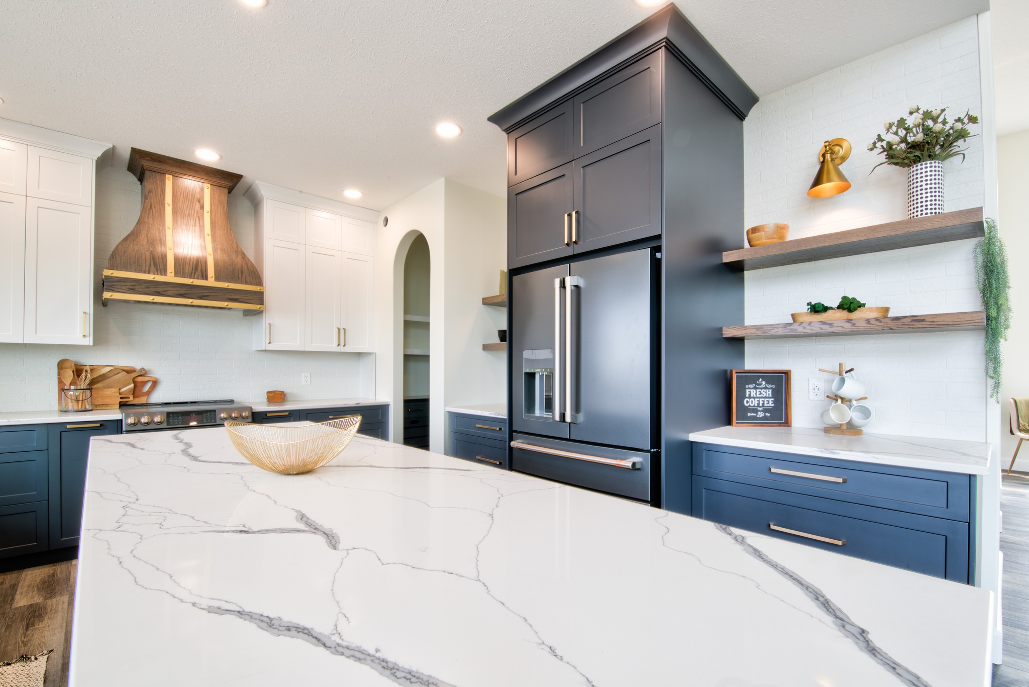 Modern kitchen with a marble island, navy and white cabinets, and stainless steel appliances. Warm lighting and decorative shelves create a cozy ambiance.